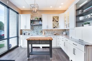 Brightly lit home kitchen with white cabinets and gray stone countertops, dark island with natural wood countertop, and chandelier lighting