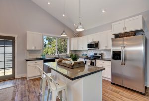 Interior of kitchen with high vaulted ceiling, pendant lights, white kitchen cabinetry, and steel appliances. 